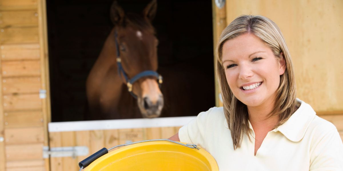 Pferdehalterin hält gelben Futtereimer, dahinter ihr Pferd in der Schweiz